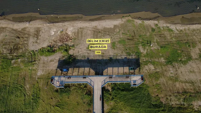 Aerial view of a waterfront pier with yellow Turkish text signs reading "IKLIM KRIZI BURADA" (Climate Crisis Here) and "350" near a sandy shoreline.