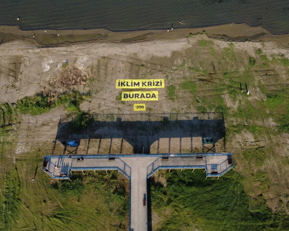 Aerial view of a waterfront pier with yellow Turkish text signs reading "IKLIM KRIZI BURADA" (Climate Crisis Here) and "350" near a sandy shoreline.
