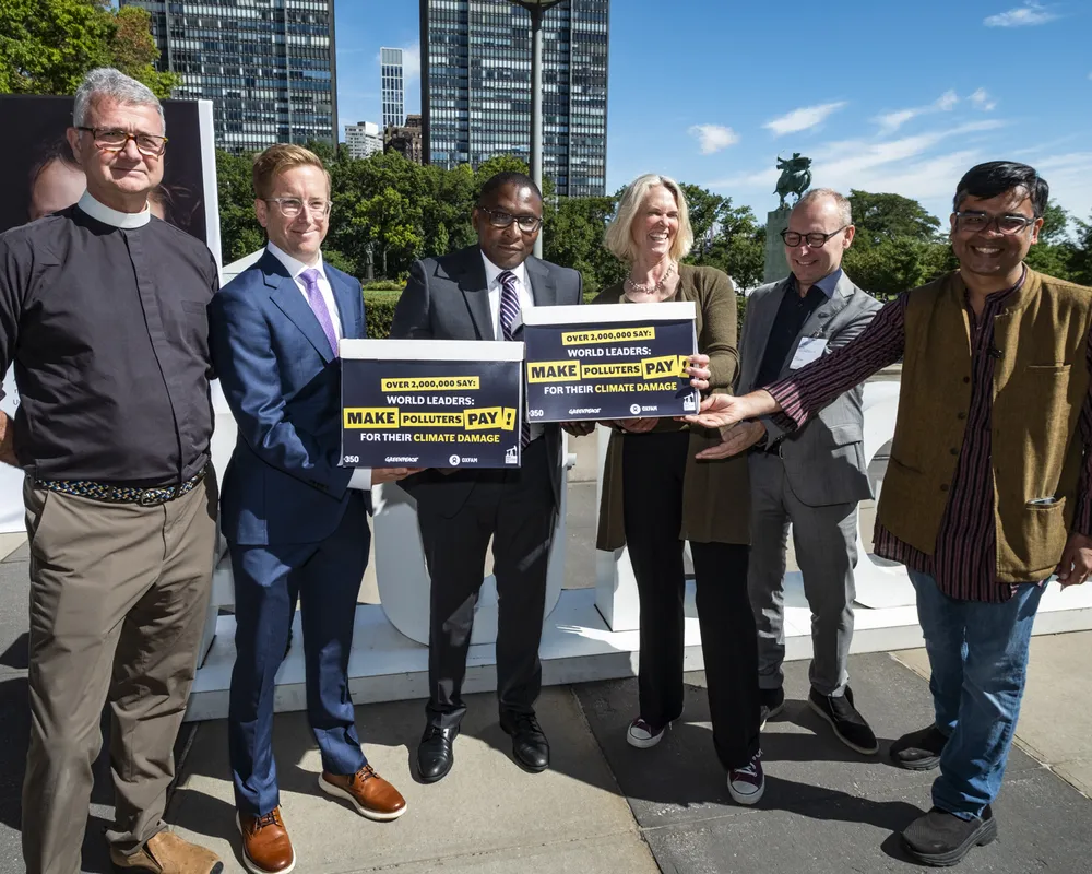 Six people standing outdoors holding signs demanding world leaders make polluters pay for their climate damage, with modern buildings and blue sky in background.