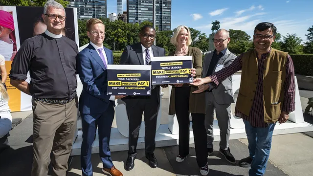 Six people standing outdoors holding signs demanding world leaders make polluters pay for their climate damage, with modern buildings and blue sky in background.