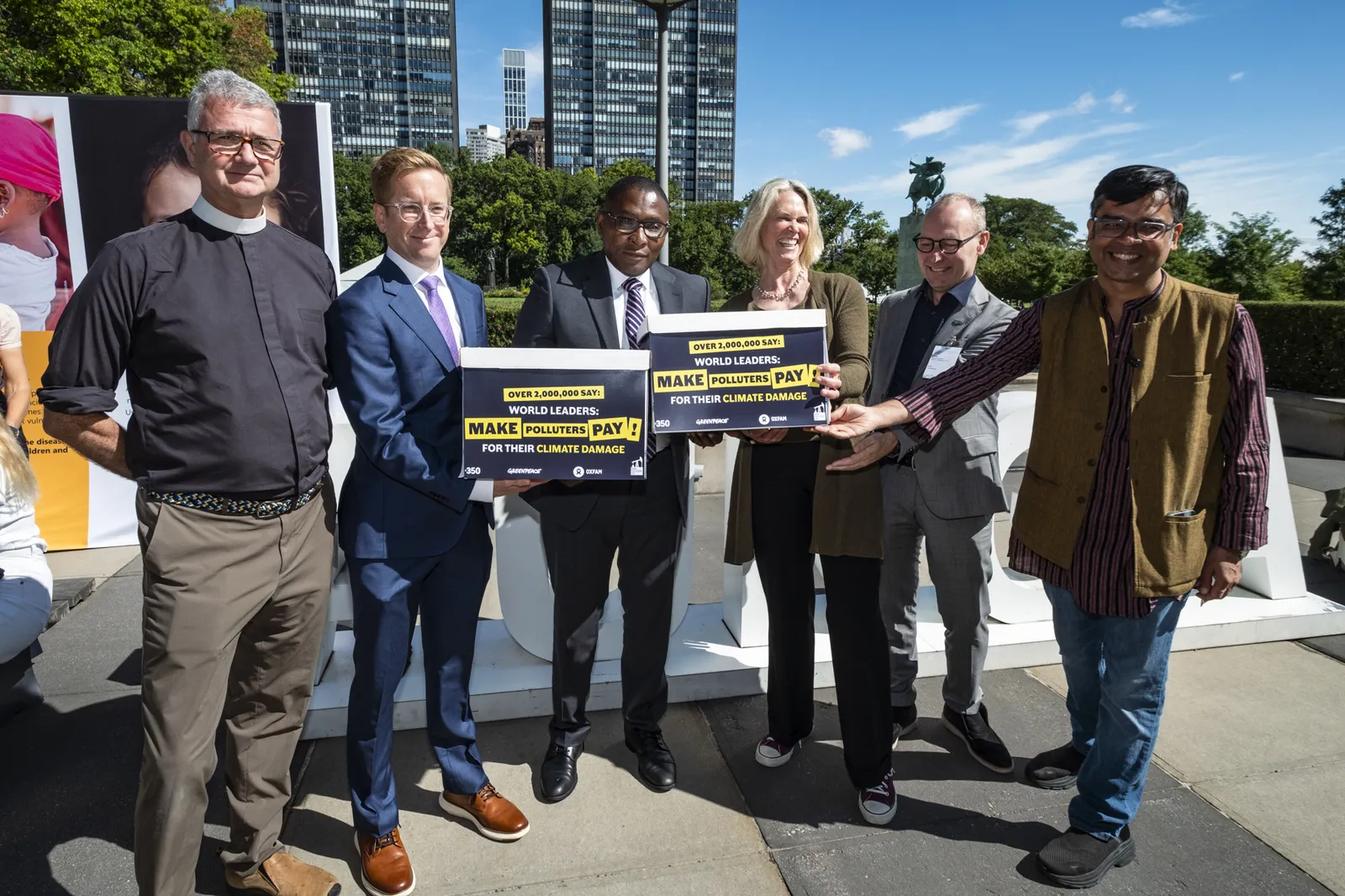 Six people standing outdoors holding signs demanding world leaders make polluters pay for their climate damage, with modern buildings and blue sky in background.