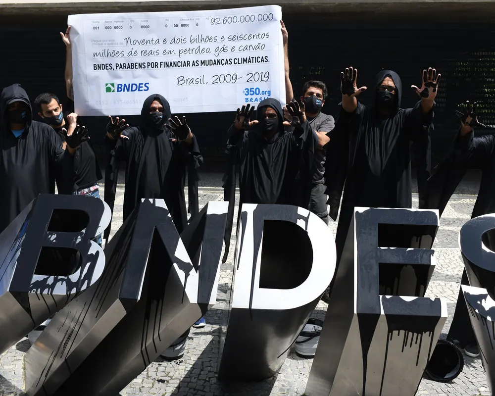 Climate activists in black robes protest at BNDES building in Brazil, holding banner about financing climate change. Large "BNDES" letters displayed in foreground during 2020-2019 demonstration.