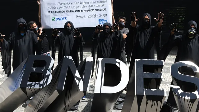 Climate activists in black robes protest at BNDES building in Brazil, holding banner about financing climate change. Large "BNDES" letters displayed in foreground during 2020-2019 demonstration.