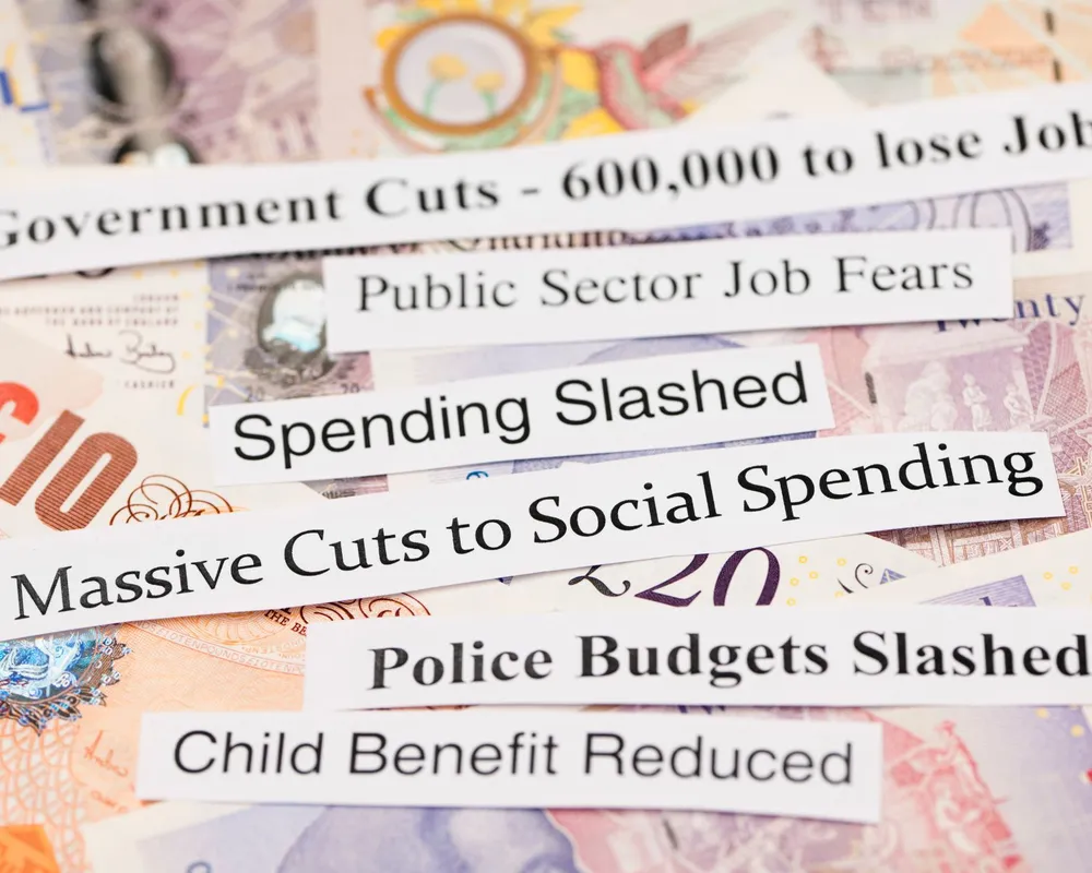 A background of money with ripped paper headlines on top. Headlines read spending slashed, massive cuts to social spending, child benefit reduced, etc.