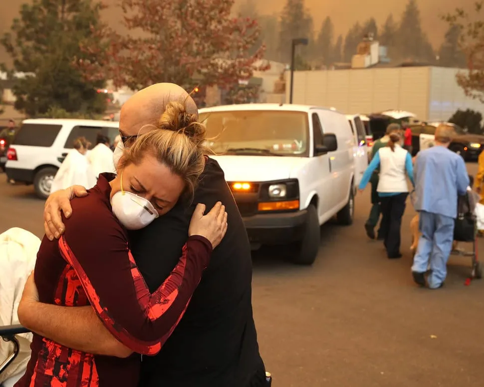 Two women embrace in a parking lot during a wildfire evacuation, with orange smoky skies overhead. Emergency vehicles and evacuees with belongings are visible in the background as people flee the disaster.