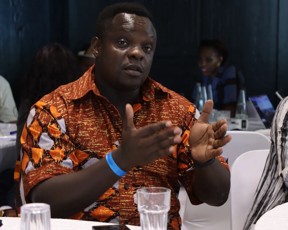 A man in an orange patterned traditional African shirt gestures while speaking at a conference table, with other attendees visible in the background.