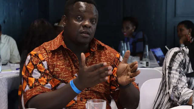 A man in an orange patterned traditional African shirt gestures while speaking at a conference table, with other attendees visible in the background.