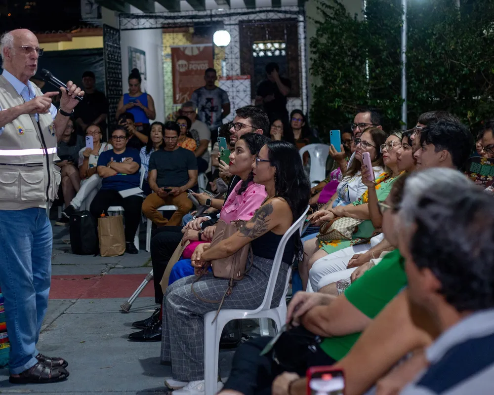 A bald man in a white vest addresses a diverse outdoor crowd seated on white plastic chairs. Attendees listen attentively, some recording on phones, with buildings and trees visible in the background.
