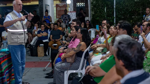 A bald man in a white vest addresses a diverse outdoor crowd seated on white plastic chairs. Attendees listen attentively, some recording on phones, with buildings and trees visible in the background.