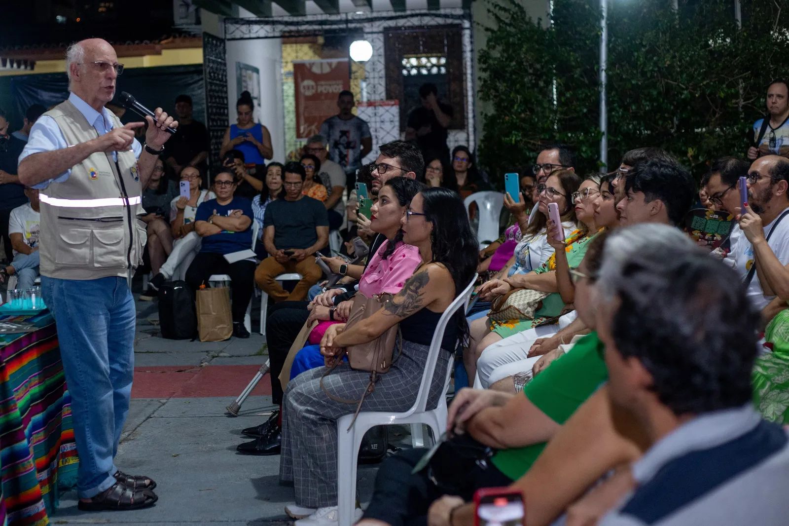 A bald man in a white vest addresses a diverse outdoor crowd seated on white plastic chairs. Attendees listen attentively, some recording on phones, with buildings and trees visible in the background.