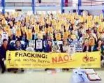 Large group photo of anti-fracking protesters holding yellow banner that reads "FRACKING KILLS" in what appears to be an indoor venue with flags visible in background. Protesters wearing yellow shirts gathered for demonstration.