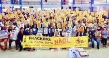 Large group photo of anti-fracking protesters holding yellow banner that reads "FRACKING KILLS" in what appears to be an indoor venue with flags visible in background. Protesters wearing yellow shirts gathered for demonstration.