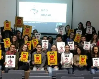 Large group of students in a classroom holding protest signs in Portuguese with "NÃO" (NO) messages. They're standing in rows with a projection screen visible in the background, appearing to participate in an organized demonstration.