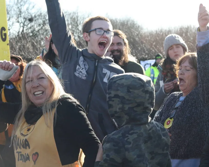 Protesters with raised fists celebrating at an outdoor demonstration, wearing casual winter clothing and holding signs, with people showing excitement and solidarity during what appears to be an environmental protest.