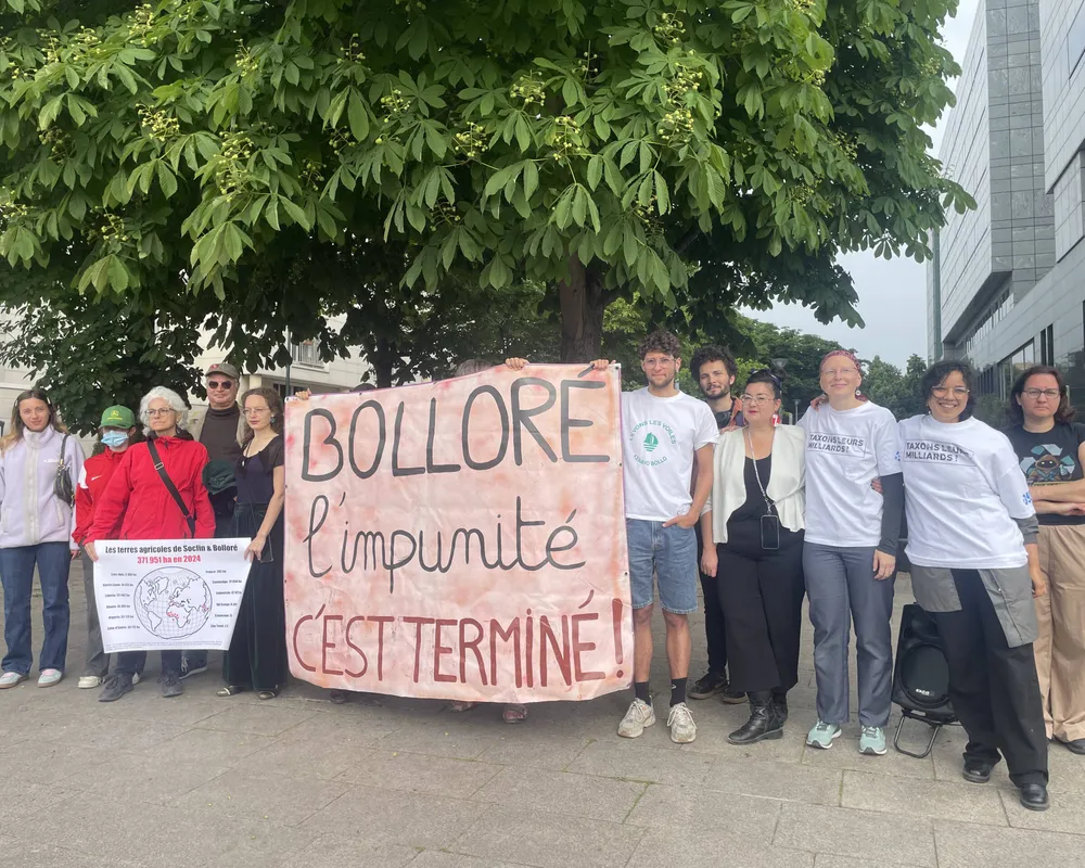 Group of protesters holding a large banner reading "BOLLORE l'impunité c'est terminé!" (Bollore impunity is over!) standing under a tree near modern buildings, advocating for corporate accountability.