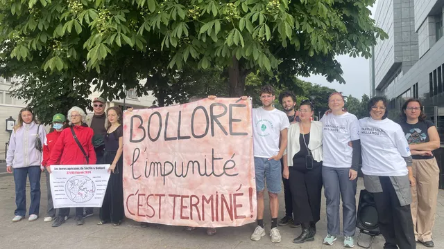 Group of protesters holding a large banner reading "BOLLORE l'impunité c'est terminé!" (Bollore impunity is over!) standing under a tree near modern buildings, advocating for corporate accountability.