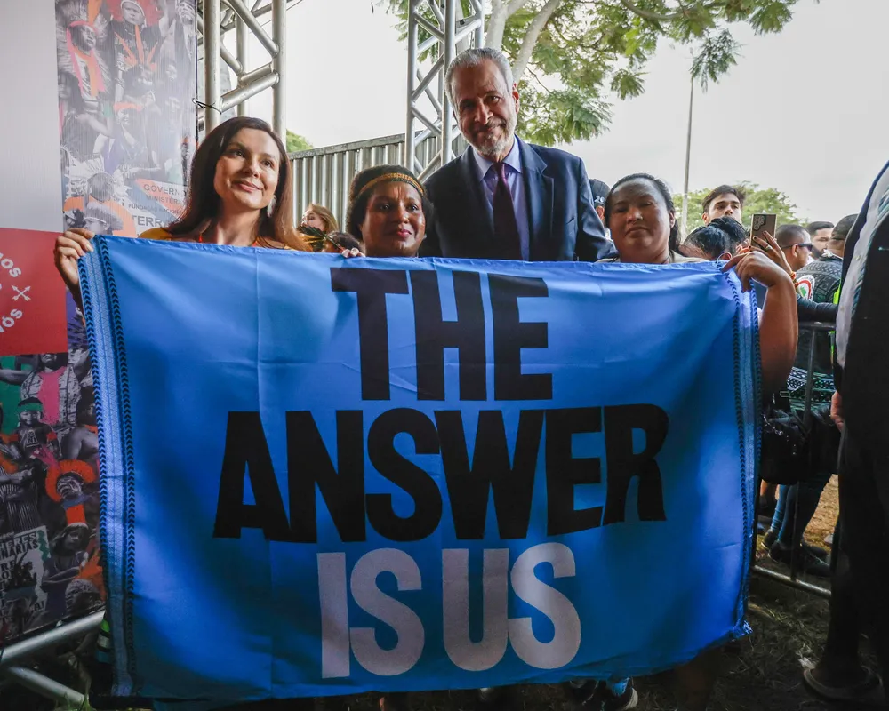 A group of people hold a blue banner reading "THE ANSWER IS US" at an outdoor event. A man in a blue suit stands with women, surrounded by attendees and metal framework visible in the background.