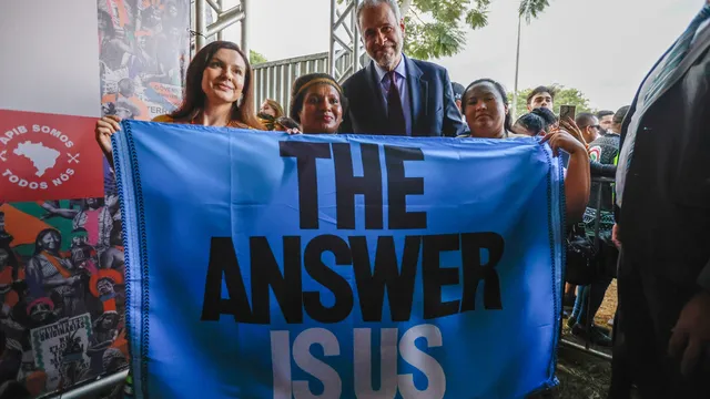 A group of people hold a blue banner reading "THE ANSWER IS US" at an outdoor event. A man in a blue suit stands with women, surrounded by attendees and metal framework visible in the background.