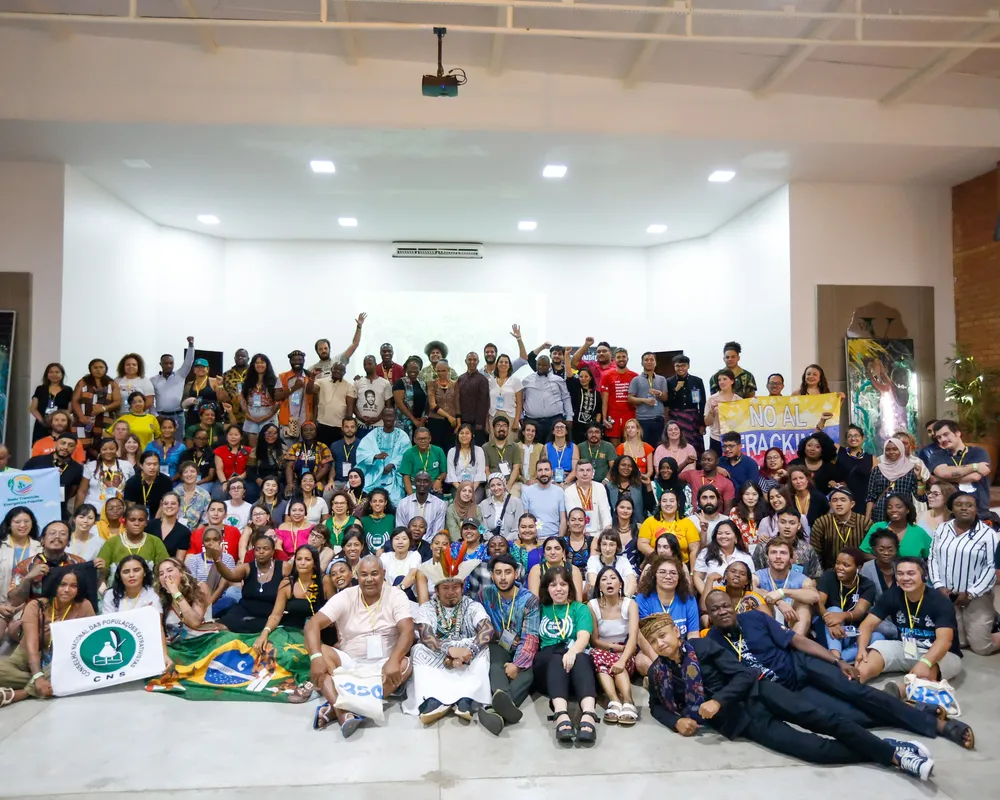 Large diverse group photo of approximately 150+ people gathered in a white-walled auditorium. Participants wear colorful clothing and hold Brazilian flags and banners, suggesting a community event or conference gathering.