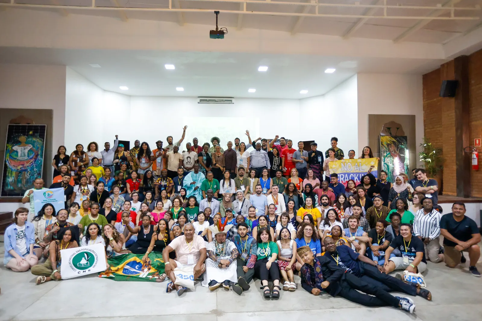 Large diverse group photo of approximately 150+ people gathered in a white-walled auditorium. Participants wear colorful clothing and hold Brazilian flags and banners, suggesting a community event or conference gathering.