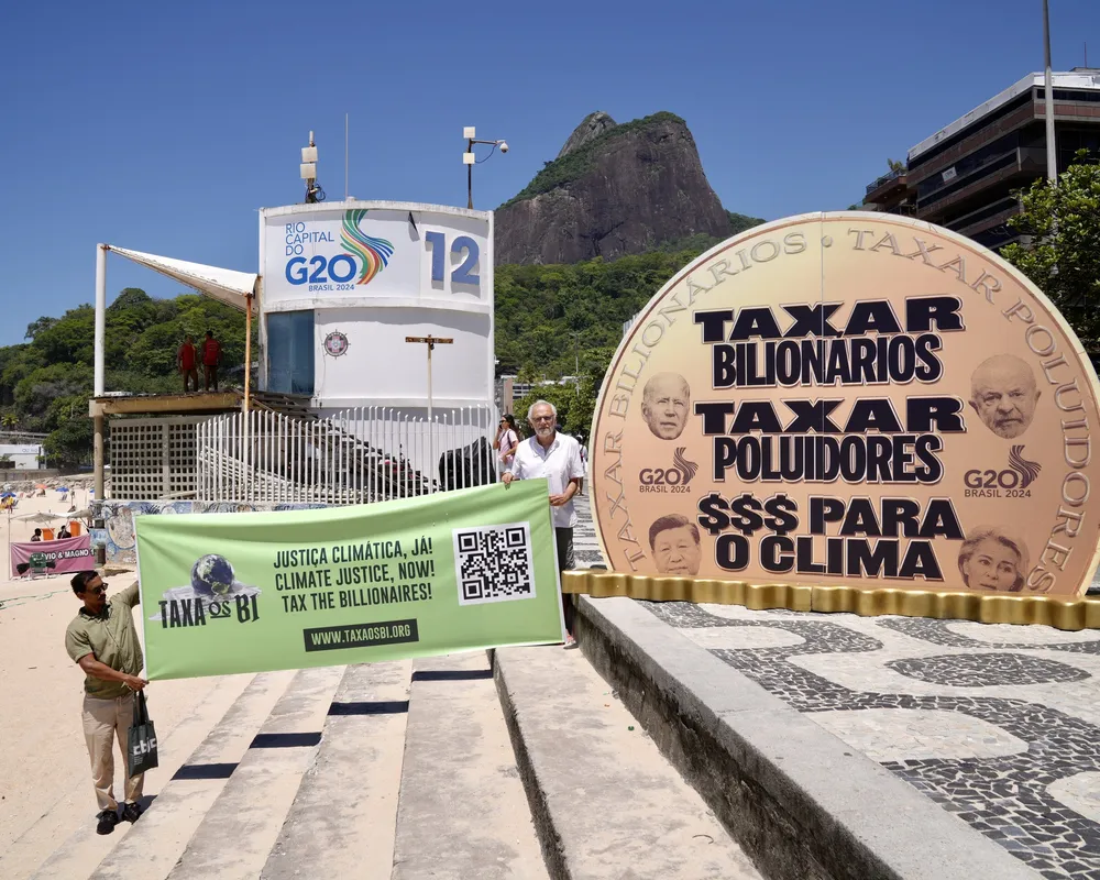 Climate activists protest at G20 summit in Rio de Janeiro, displaying a large coin prop with Portuguese text demanding billionaires pay taxes for climate action, with beach and mountain backdrop.