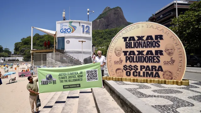Climate activists protest at G20 summit in Rio de Janeiro, displaying a large coin prop with Portuguese text demanding billionaires pay taxes for climate action, with beach and mountain backdrop.