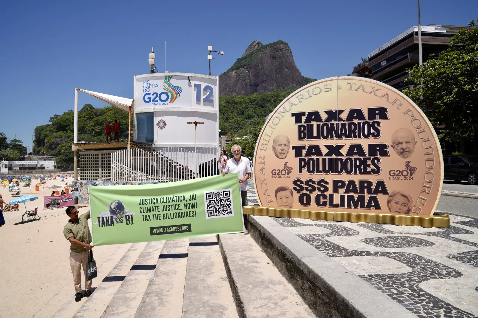 Climate activists protest at G20 summit in Rio de Janeiro, displaying a large coin prop with Portuguese text demanding billionaires pay taxes for climate action, with beach and mountain backdrop.