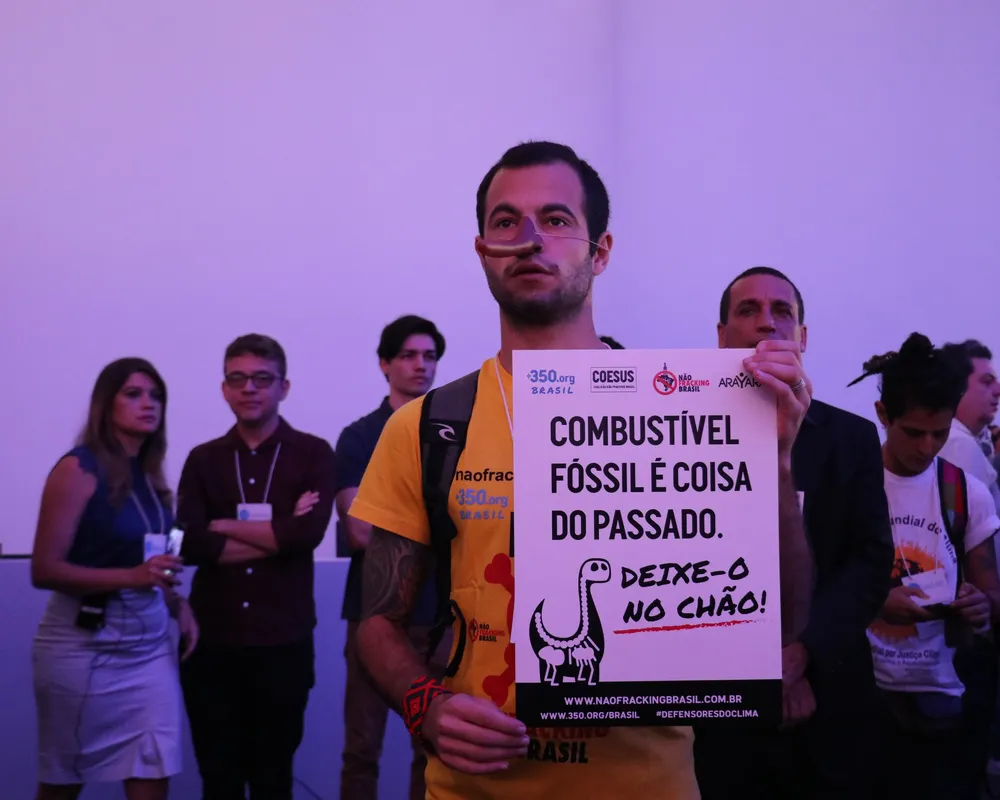 Climate activists protest at event holding sign that reads "Fossil fuel is a thing of the past. Leave it in the ground!" in Portuguese, with supporters standing behind in purple-lit room.