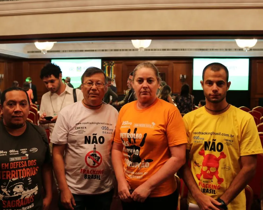 Four activists wearing protest t-shirts with "NÃO" (NO) messages stand together at a conference venue in Brazil, advocating against fracking and in defense of their territories.
