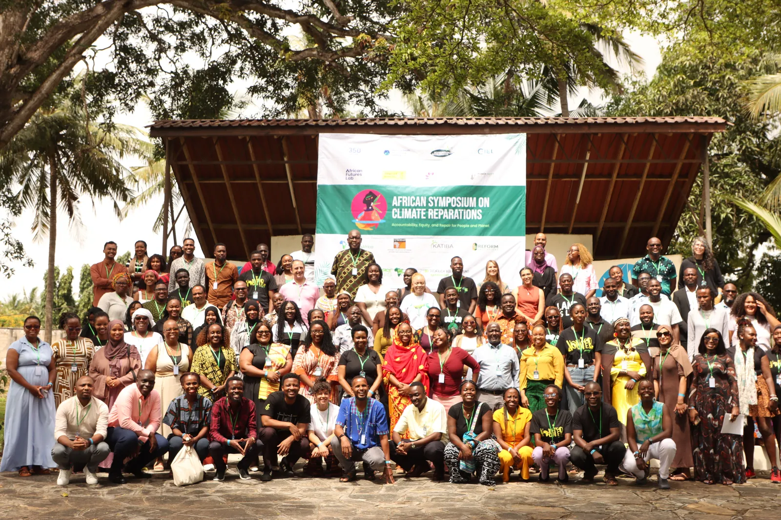 Large group photo at the African Symposium on Climate Reparations. Diverse attendees in colorful attire pose outdoors under a covered pavilion with palm trees and tropical vegetation in the background.