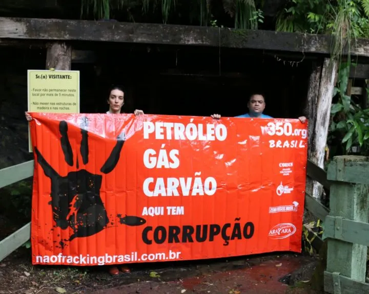 Two activists hold a large orange protest banner reading "PETRÓLEO GÁS CARVÃO AQUI TEM CORRUPÇÃO" against a weathered wooden structure surrounded by lush greenery.