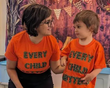 Two people wearing matching bright orange t-shirts with colorful lettering that reads "EVERY CHILD" and "EVERY CHILD MATTERS" stand together smiling at each other against a decorative background with bunting.