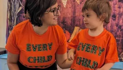 Two people wearing matching bright orange t-shirts with colorful lettering that reads "EVERY CHILD" and "EVERY CHILD MATTERS" stand together smiling at each other against a decorative background with bunting.
