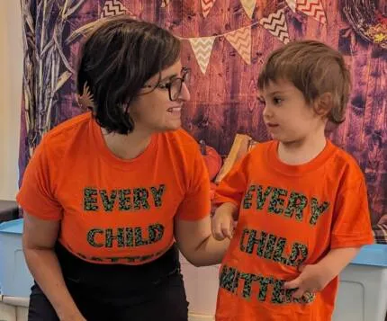 Two people wearing matching bright orange t-shirts with colorful lettering that reads "EVERY CHILD" and "EVERY CHILD MATTERS" stand together smiling at each other against a decorative background with bunting.