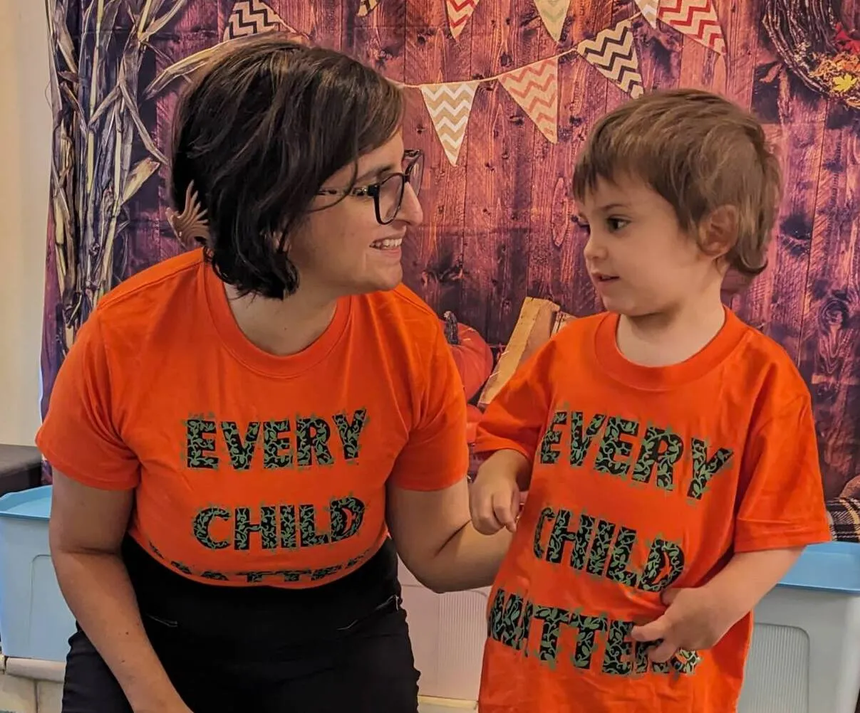 A woman and child wear matching orange shirts with "EVERY CHILD MATTERS" text, standing together indoors against a decorative backdrop with bunting flags, sharing a warm moment while looking at each other and smiling.