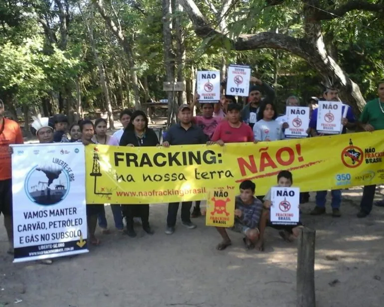 Community members protest against fracking in Brazil, holding a large yellow banner reading "FRACKING: na nossa NÃO!" and various signs opposing the practice, gathered in a tree-lined outdoor area.
