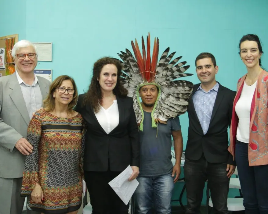 Six people pose together against a turquoise wall. The central figure wears a traditional feathered headdress. The group appears diverse, dressed in business and casual attire, suggesting a formal meeting or cultural event.
