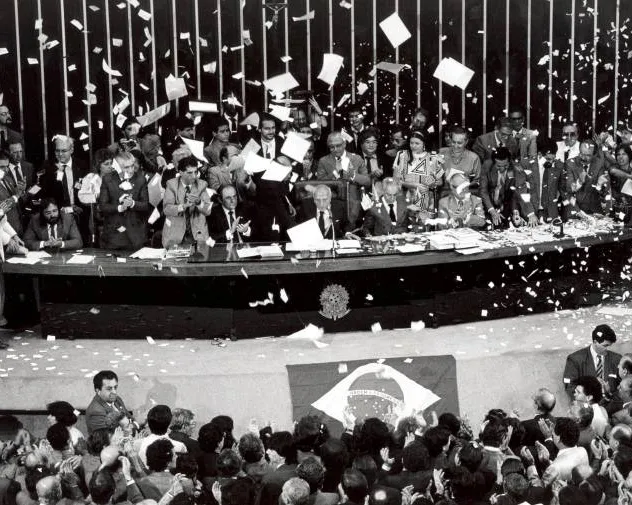 Imagem em preto e branco, Ulysses Guimarães com parlamentares festejam em meio a papéis picados. pronunciou as palavras históricas: "Declaro promulgado o documento da liberdade, da democracia e da justiça social do Brasil"