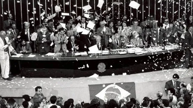 Imagem em preto e branco, Ulysses Guimarães com parlamentares festejam em meio a papéis picados. pronunciou as palavras históricas: "Declaro promulgado o documento da liberdade, da democracia e da justiça social do Brasil"