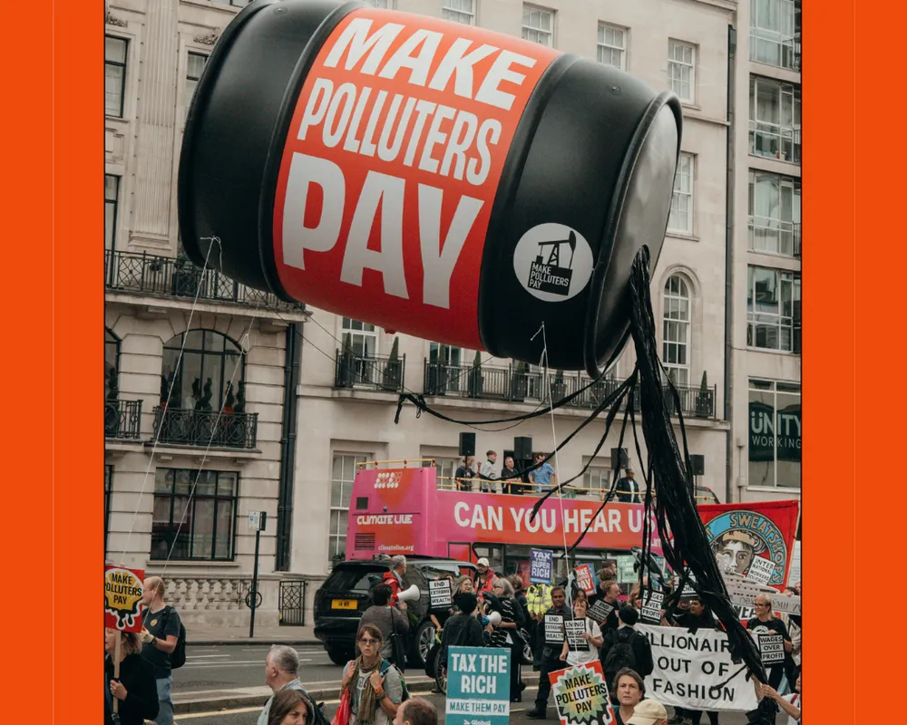 A protest scene featuring a giant inflatable barrel reading "MAKE POLLUTERS PAY" suspended above demonstrators holding signs about fossil fuels and climate action in an urban setting.