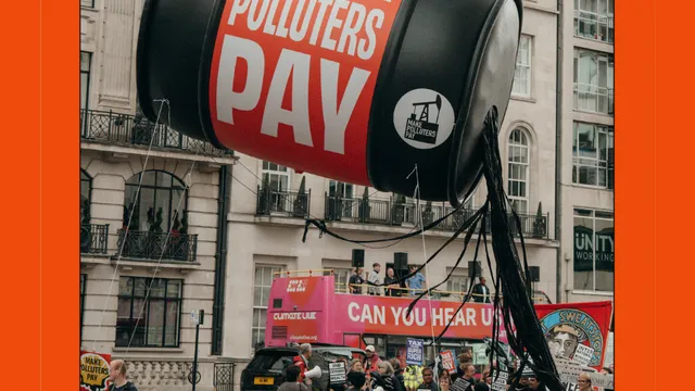 A protest scene featuring a giant inflatable barrel reading "MAKE POLLUTERS PAY" suspended above demonstrators holding signs about fossil fuels and climate action in an urban setting.