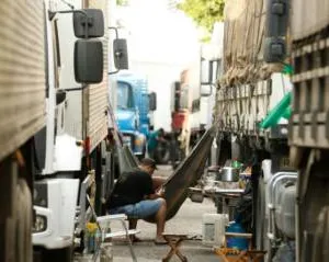 A person relaxes in a hammock strung between two large trucks on a narrow urban street, creating an improvised resting spot amid the congested traffic and industrial surroundings.