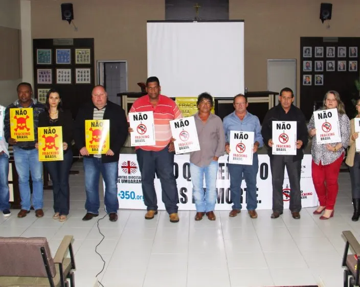 A group of people stand in a meeting room holding yellow and white protest signs with "NÃO" (NO in Portuguese), advocating against something. Signs visible on floor and held by participants in casual attire.