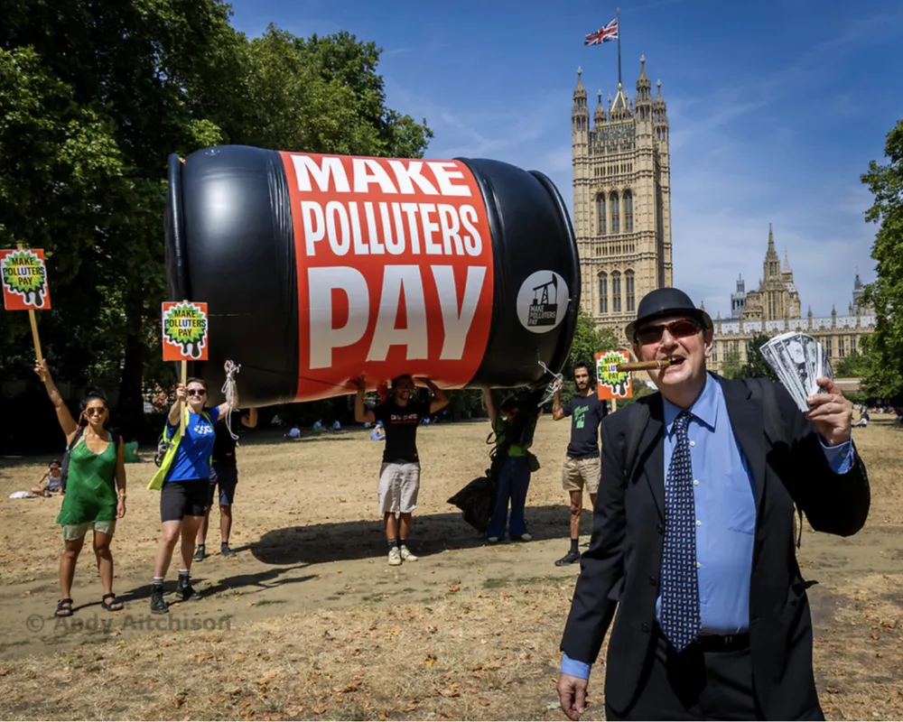 Environmental protesters with giant inflatable oil barrel reading "MAKE POLLUTERS PAY" demonstrate near Parliament in London. Activists hold signs demanding climate action while a suited figure poses in foreground.