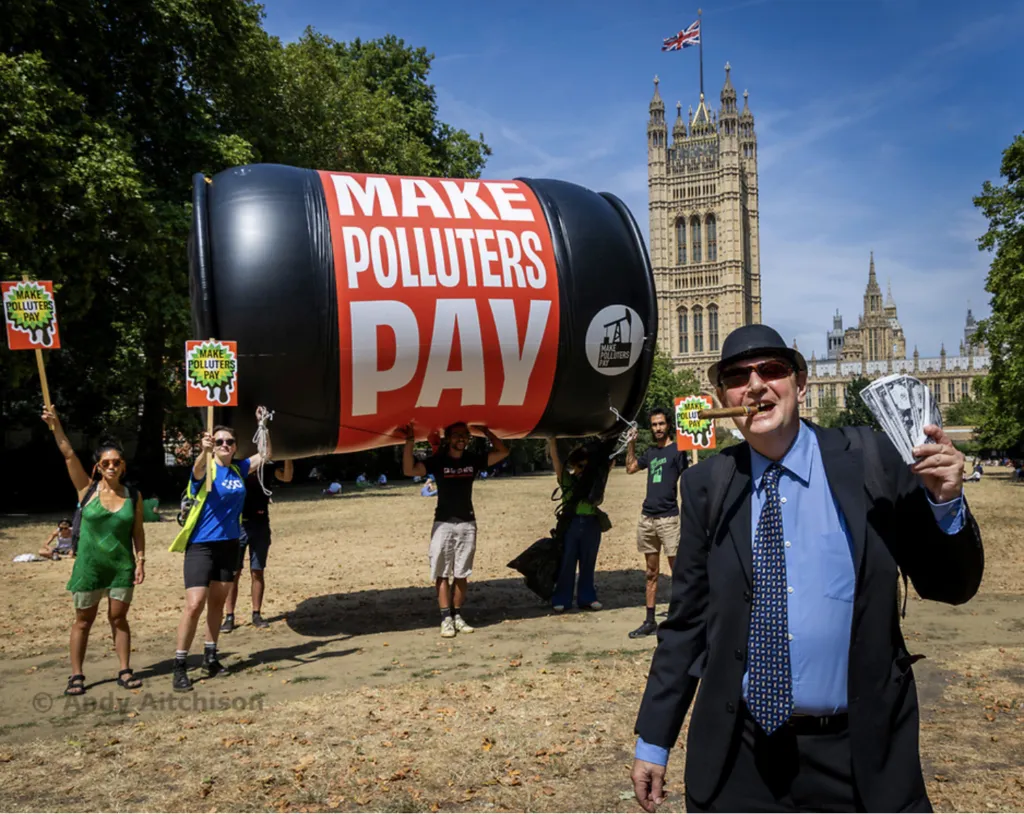 Environmental protesters with giant inflatable oil barrel reading "MAKE POLLUTERS PAY" demonstrate near Parliament in London. Activists hold signs demanding climate action while a suited figure poses in foreground.
