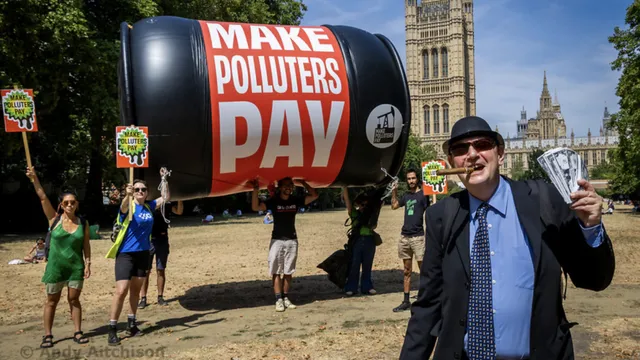 Environmental protesters with giant inflatable oil barrel reading "MAKE POLLUTERS PAY" demonstrate near Parliament in London. Activists hold signs demanding climate action while a suited figure poses in foreground.