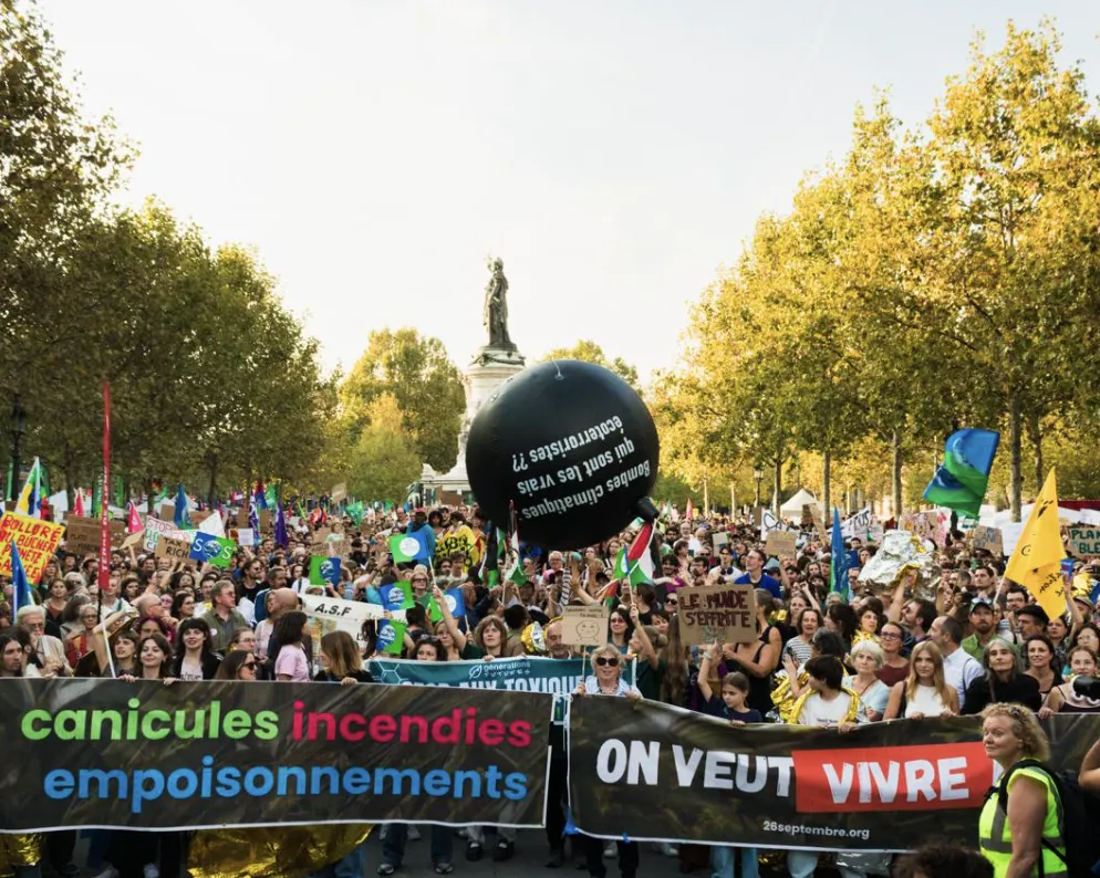 Large climate protest march with crowds holding banners reading "canicules incendies empoisonnements" and "ON VEUT VIVRE" among trees in a public square, with a large black balloon and statue visible in background.