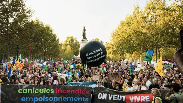 Large climate protest march with crowds holding banners reading "canicules incendies empoisonnements" and "ON VEUT VIVRE" among trees in a public square, with a large black balloon and statue visible in background.