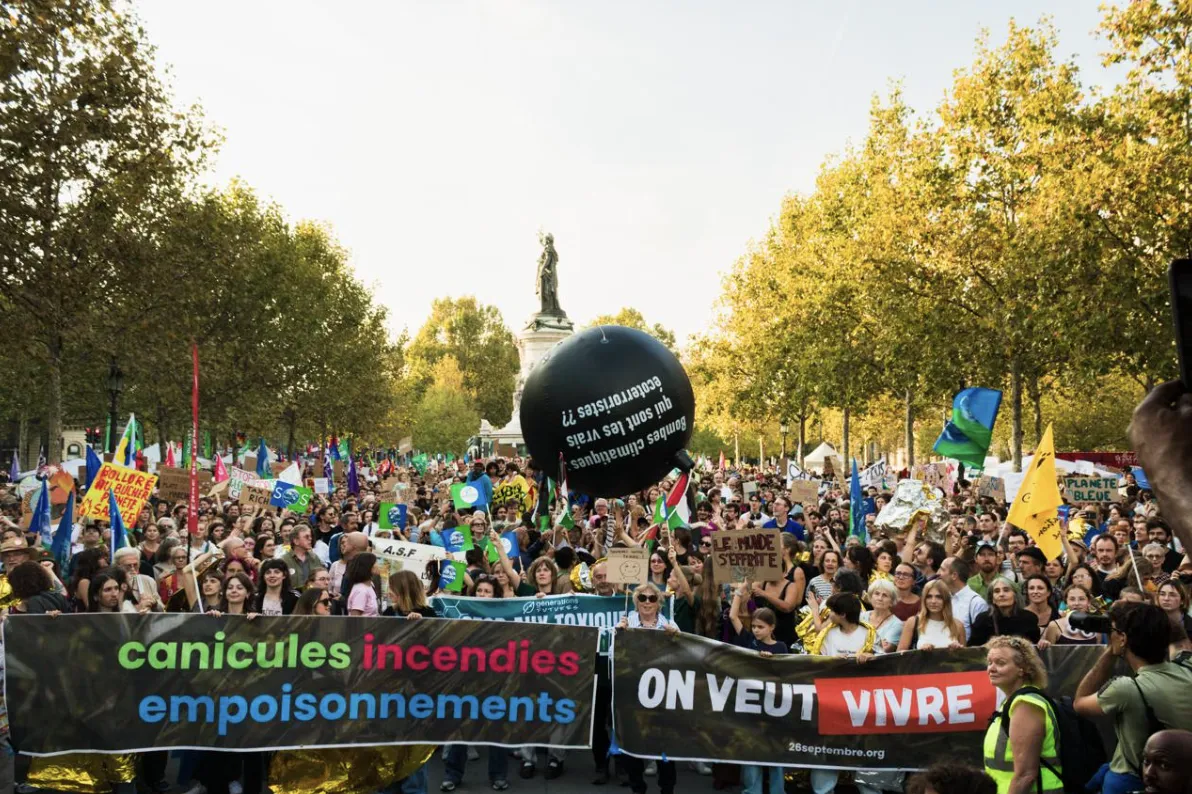 Large climate protest march with crowds holding banners reading "canicules incendies empoisonnements" and "ON VEUT VIVRE" among trees in a public square, with a large black balloon and statue visible in background.