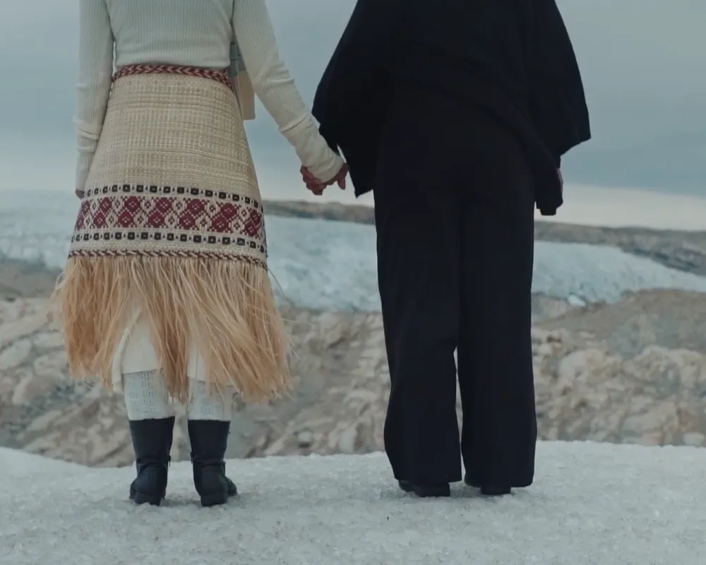 Two women hold hands in solidarity, standing side by side at the edge of a glacier, backs to camera.